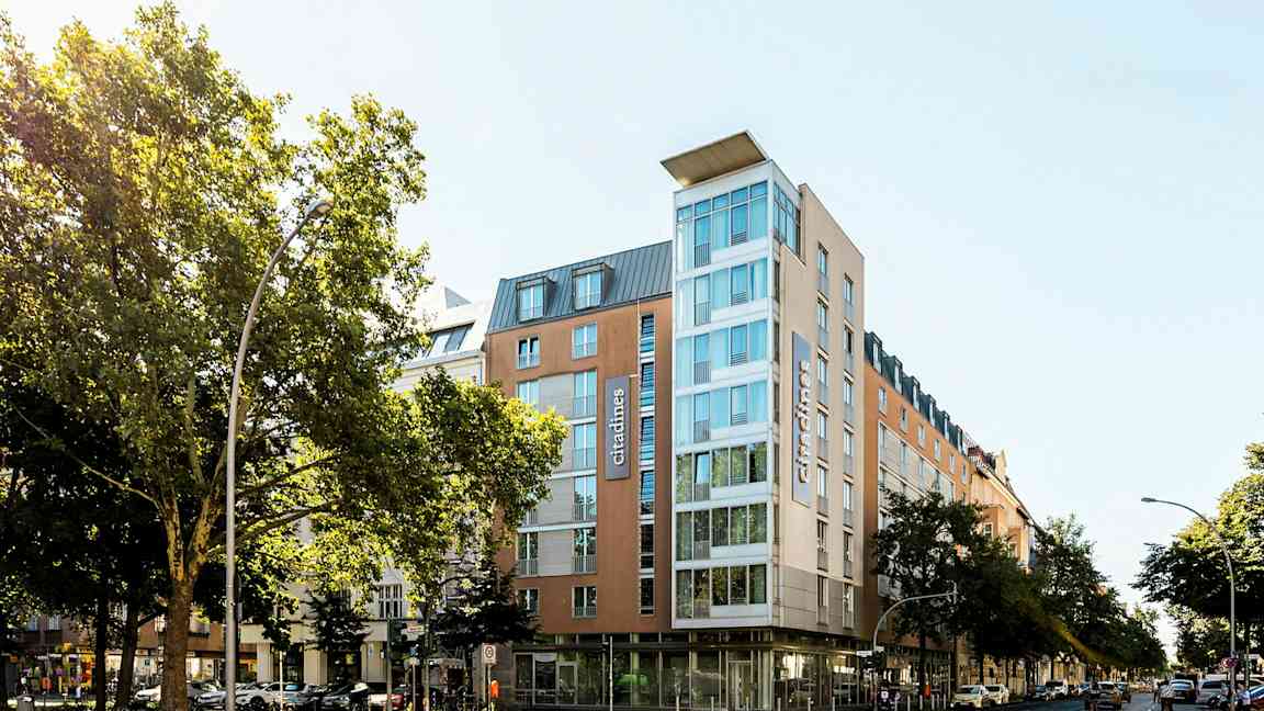 Et moderne hotel med glasfacade omgivet af træer og bymiljø. (AI-genereret tekst)