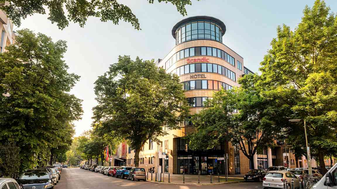 En byudsigt med facaden af et Scandic-hotel omgivet af træer og parkerede biler. (AI-genereret tekst)