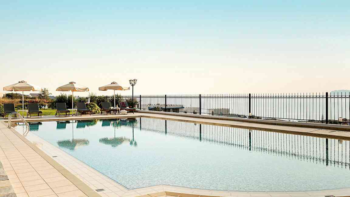 En udendørs pool med liggestole og parasoller langs kanten, med havet i baggrunden. (AI-genereret tekst)