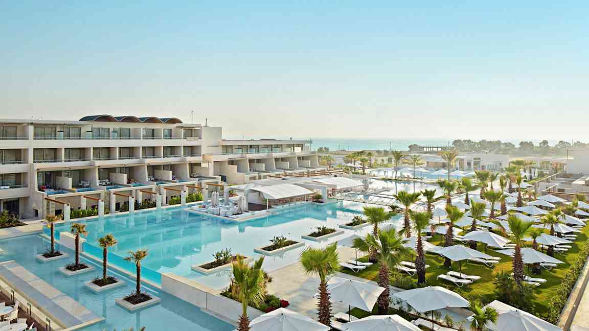 Et hotelområde med flere bygninger, en stor swimmingpool og omkringliggende grønne områder fyldt med parasoller og liggestole. I baggrunden ses havet. (AI-genereret tekst)