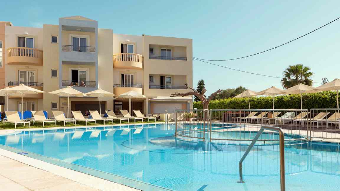 Billede af et hotelområde med en pool, liggestole og parasoller. I baggrunden ses en bygning med altaner. (AI-genereret tekst)