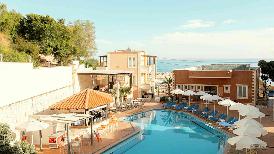 En hotelpool med liggestole og parasoller ved siden af samt bygninger og vegetation i baggrunden. (AI-genereret tekst)