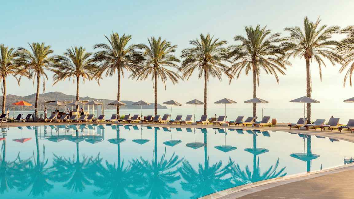 En pool med liggestole og parasoller, omgivet af palmer. Havet kan ses i baggrunden. (AI-genereret tekst)