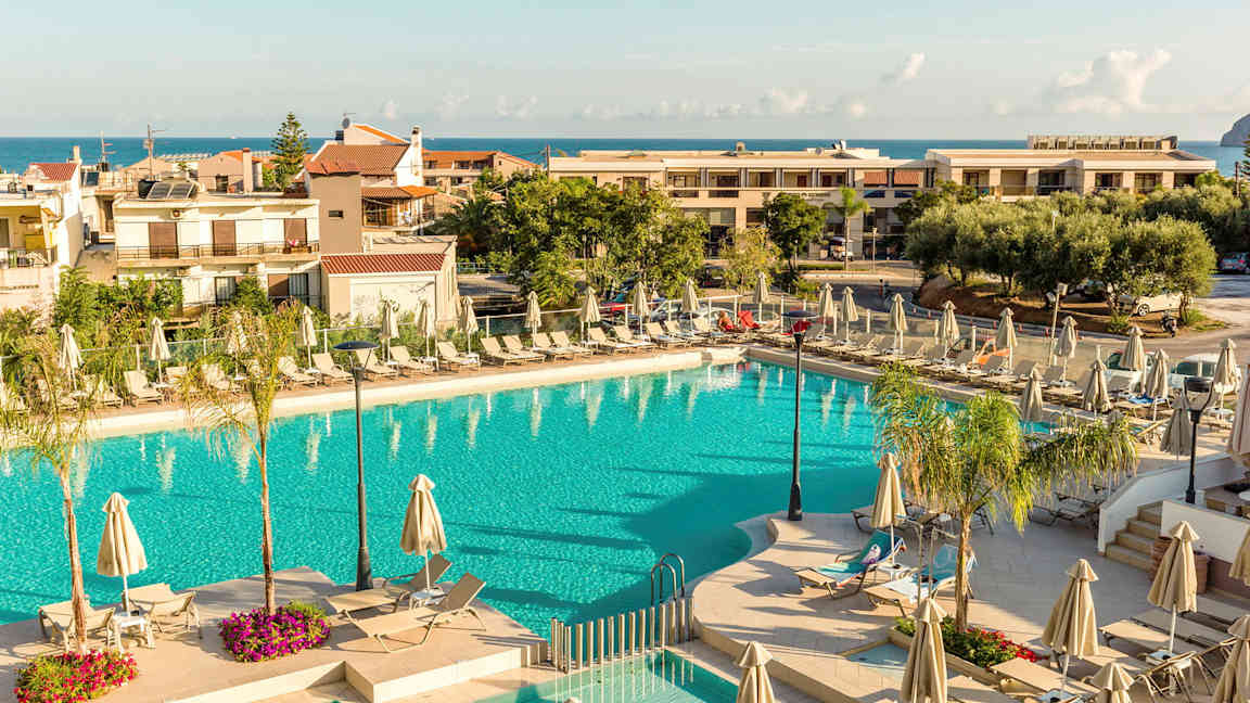 En udendørs swimmingpool omgivet af solsenge og parasoller med bygninger og havet i baggrunden. (AI-genereret tekst)