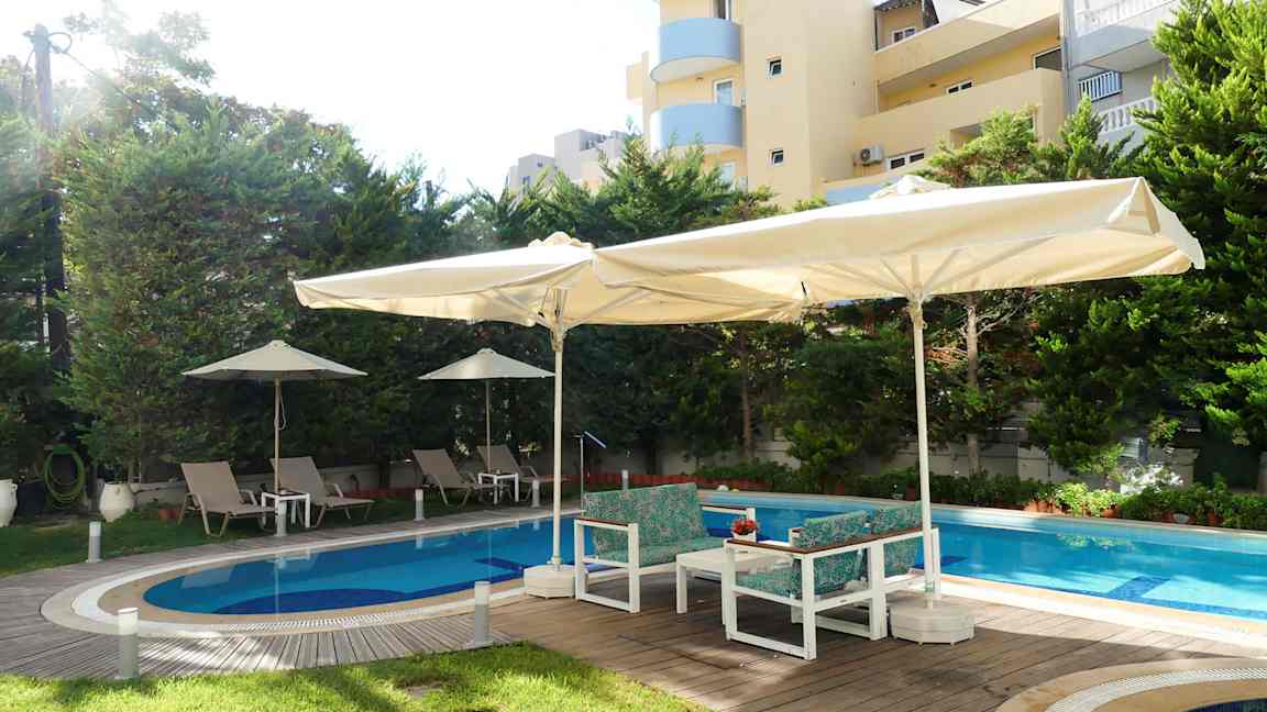 Udendørs pool med liggestole og parasoller, omgivet af træer og bygninger. (AI-genereret tekst)