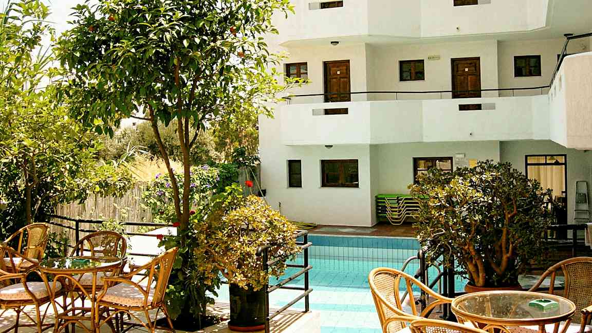 En udendørs pool omgivet af beplantning og siddepladser samt en hvid bygning med altaner i baggrunden. (AI-genereret tekst)