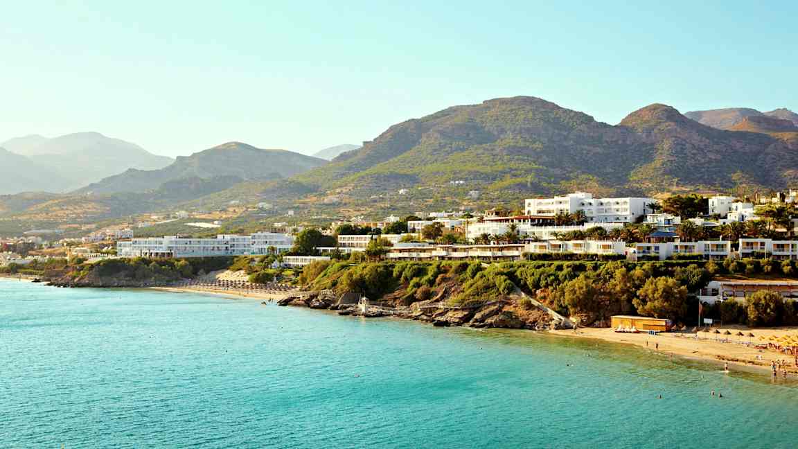 Flere bygninger nær en kyst med blåt hav og bjerge i baggrunden. (AI-genereret tekst)