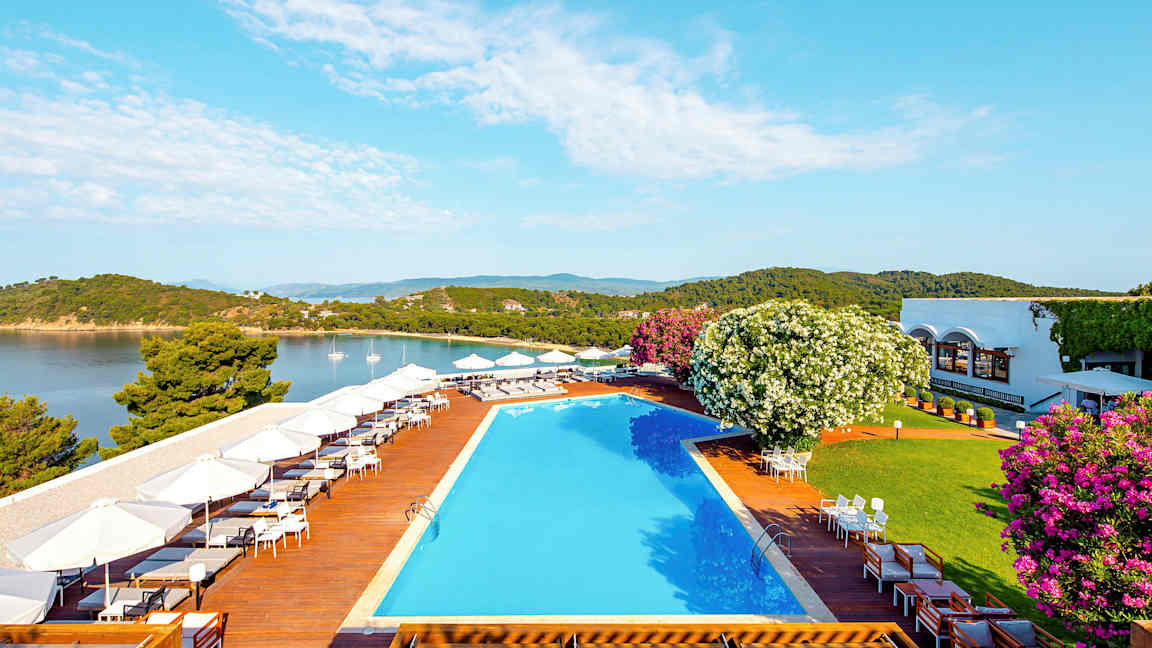 Udendørs område med pool og terrasse ved havet, omgivet af frodig vegetation og bygninger i baggrunden. (AI-genereret tekst)