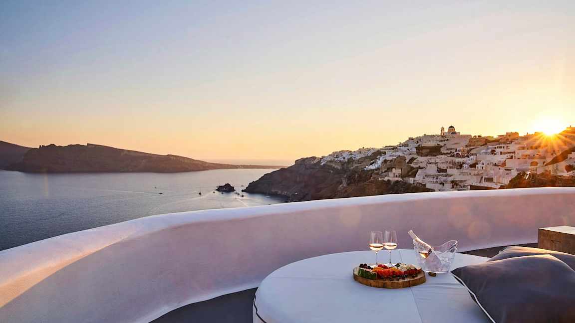 Udsigt over en solnedgang ved havet med hvide bygninger på en klippe og en terrasse med bord, mad og drikke i forgrunden. (AI-genereret tekst)