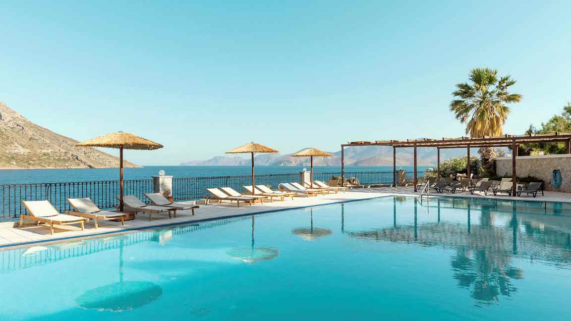 En udendørs pool med liggestole og parasoller ved siden af, samt havudsigt i baggrunden. (AI-genereret tekst)