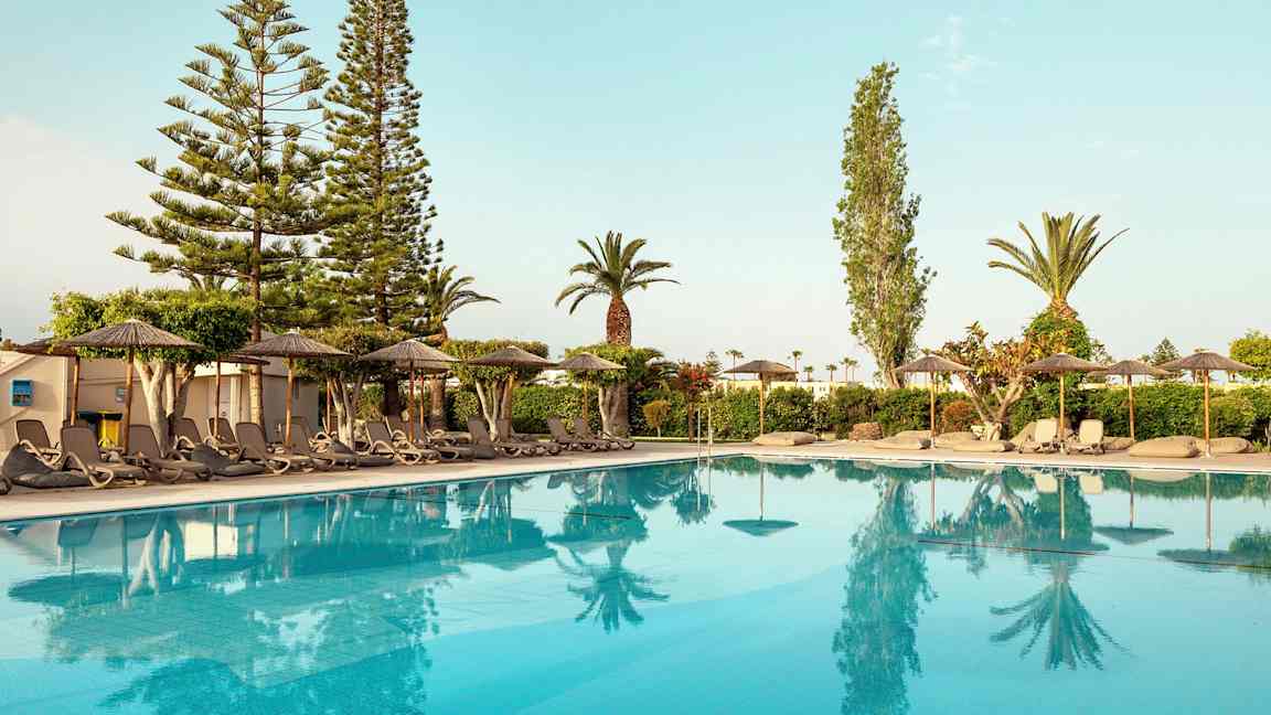 Udsigt over en pool med liggestole og parasoller ved kanten, omgivet af træer og buske. (AI-genereret tekst)