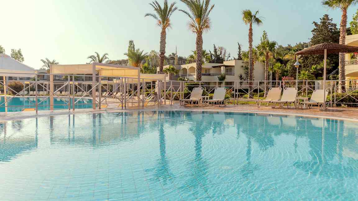 Poolområde med liggestole og parasoller, omgivet af palmer og bygninger. (AI-genereret tekst)