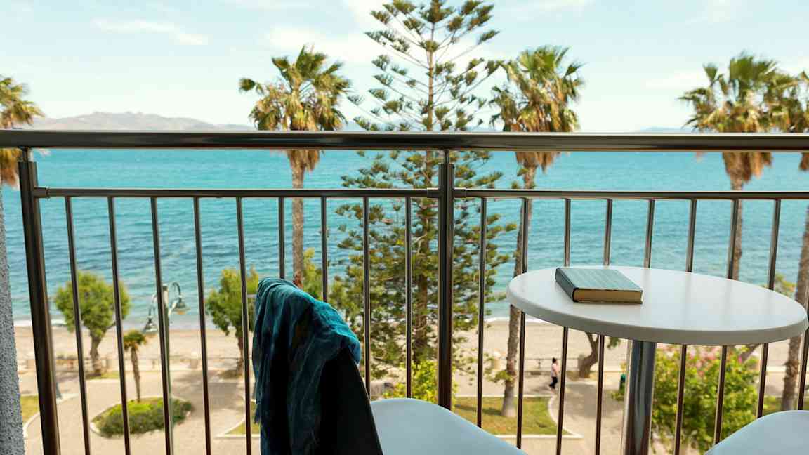 Udsigt fra en altan med bord og stole mod havet og palmer. (AI-genereret tekst)