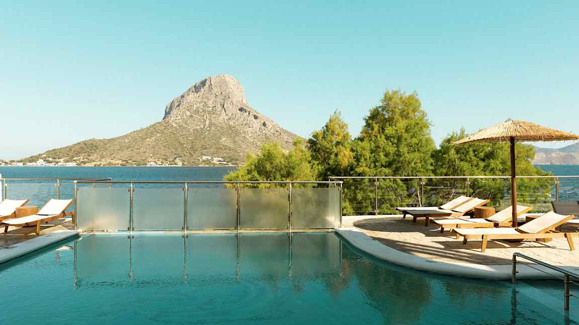En udendørs pool med liggestole og parasol, beliggende tæt på havet med en bakkelignende høj i baggrunden. (AI-genereret tekst)