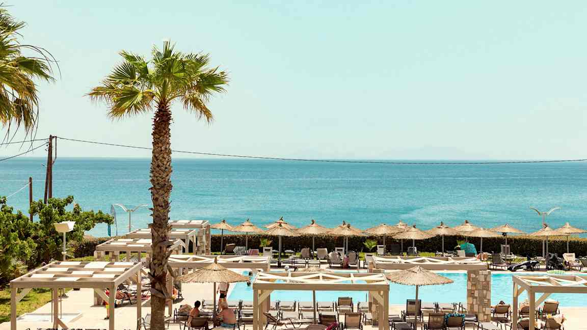 En udsigt over udendørs poolområde med liggestole, parasoller og havet i baggrunden. (AI-genereret tekst)