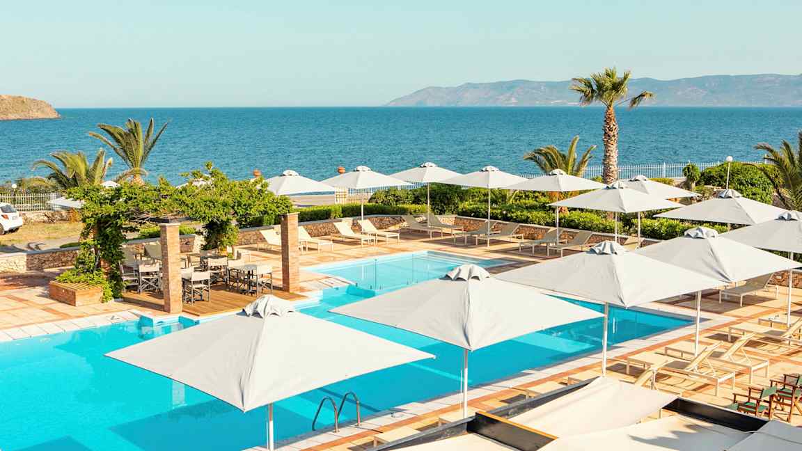 En pool med liggestole og parasoller, udsigt til havet med vegetation omkring. (AI-genereret tekst)