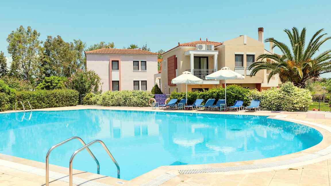 En pool omgivet af liggestole og parasoller, med bygninger og grønt i baggrunden. (AI-genereret tekst)