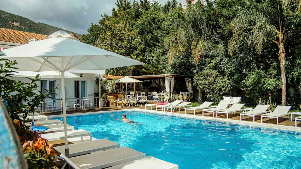 En udendørs pool omgivet af hvide liggestole og parasoller nær en bygning og grønne områder. (AI-genereret tekst)