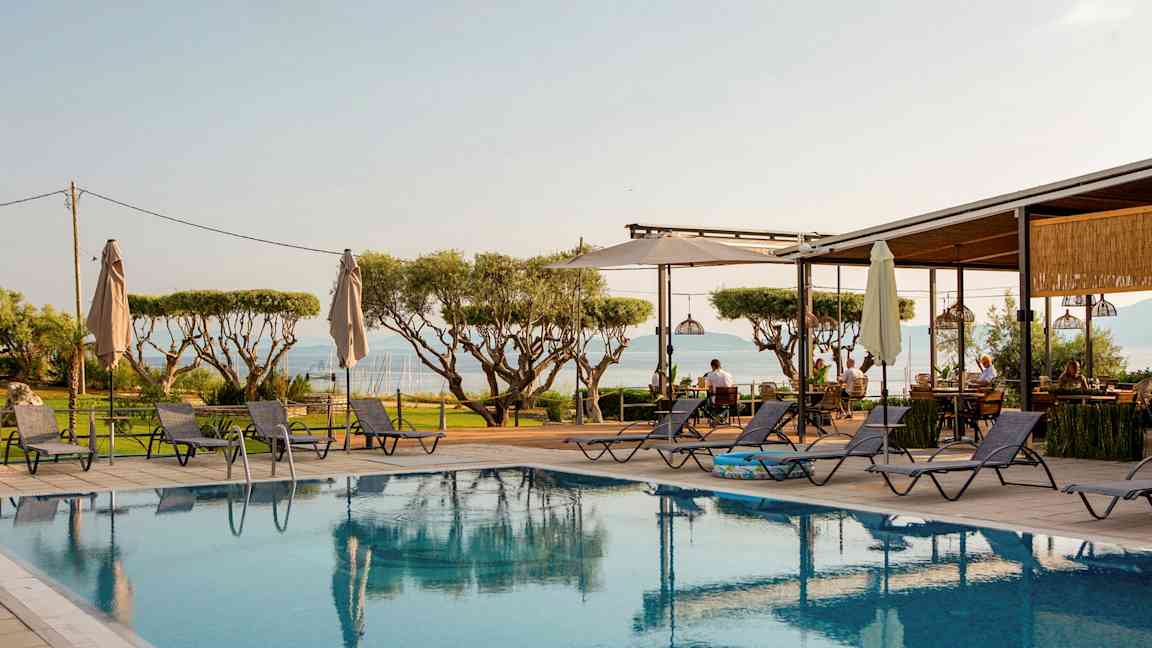 Udsigt over en pool med liggestole omkring, træer i baggrunden og havet i det fjerne. (AI-genereret tekst)