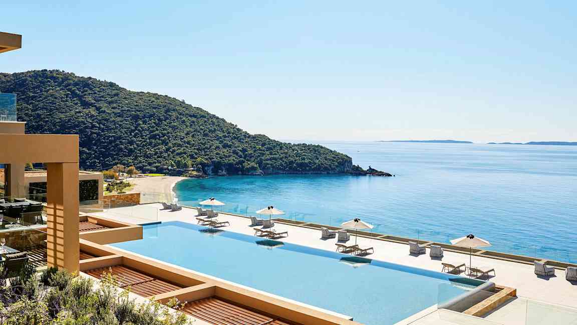 Udsigt over en pool med solsenge og parasoller, en kystlinje og hav i baggrunden. (AI-genereret tekst)