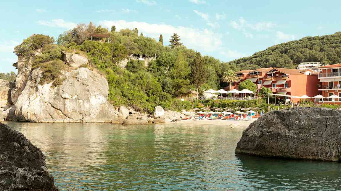 Billede af en klippestrand med parasoller og liggestole samt bygninger omgivet af grønt. (AI-genereret tekst)