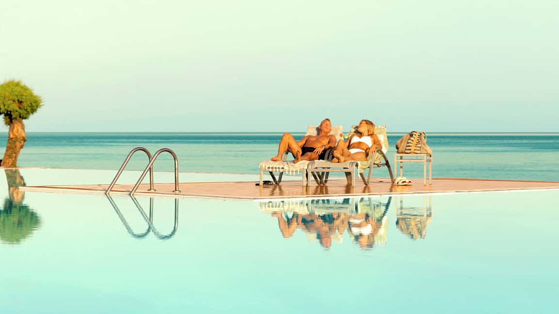 To personer ligger på liggestole ved en pool med udsigt over havet. (AI-genereret tekst)