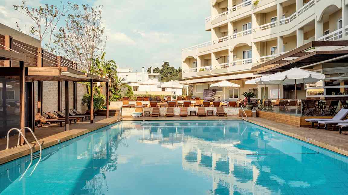 En udendørs swimmingpool med liggestole og parasoller ved siden af og et fler-etagers hotel i baggrunden. (AI-genereret tekst)
