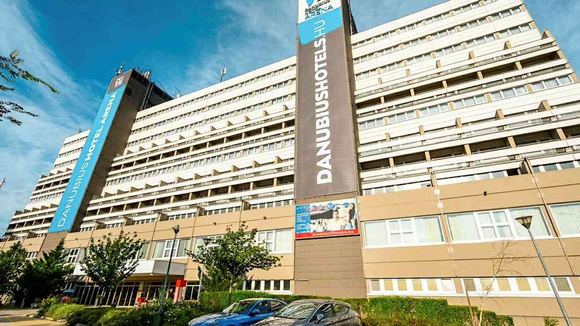 En stor hotelbygning med flere etager med skiltet ”Danubius Hotel Arena” synligt. Foran bygningen er parkerede biler og nogle små træer. (AI-genereret tekst)