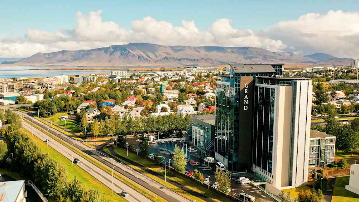 Et bymiljø med en motorvej i forgrunden, træer, bygninger og et større hotel. I baggrunden ses et bjerglandskab. (AI-genereret tekst)