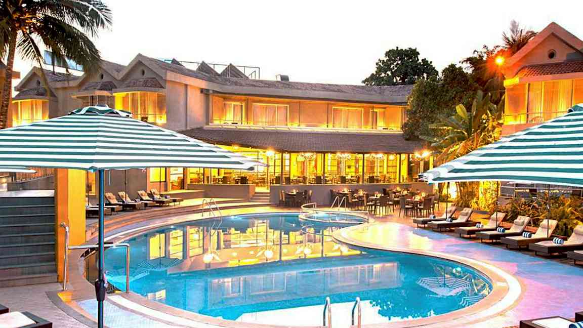 Et hotel med et oplyst poolområde med liggestole og parasoller foran bygningen. (AI-genereret tekst)