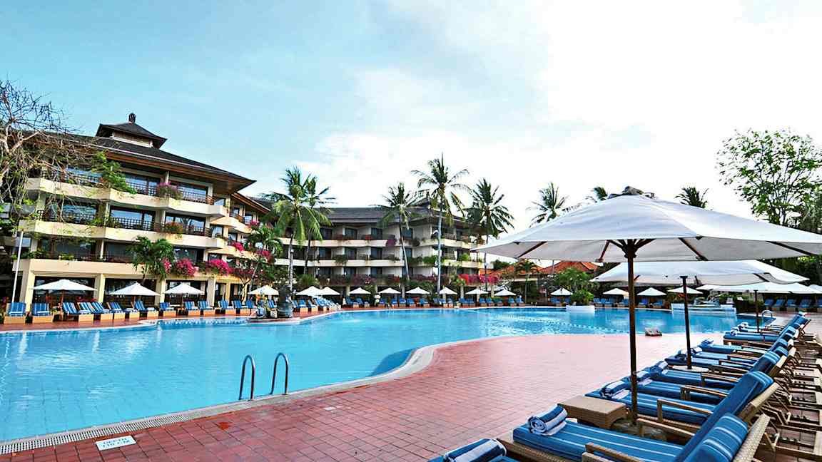 Billede af en udendørs pool omgivet af liggestole og parasoller med et hotel i flere etager og palmer i baggrunden. (AI-genereret tekst)