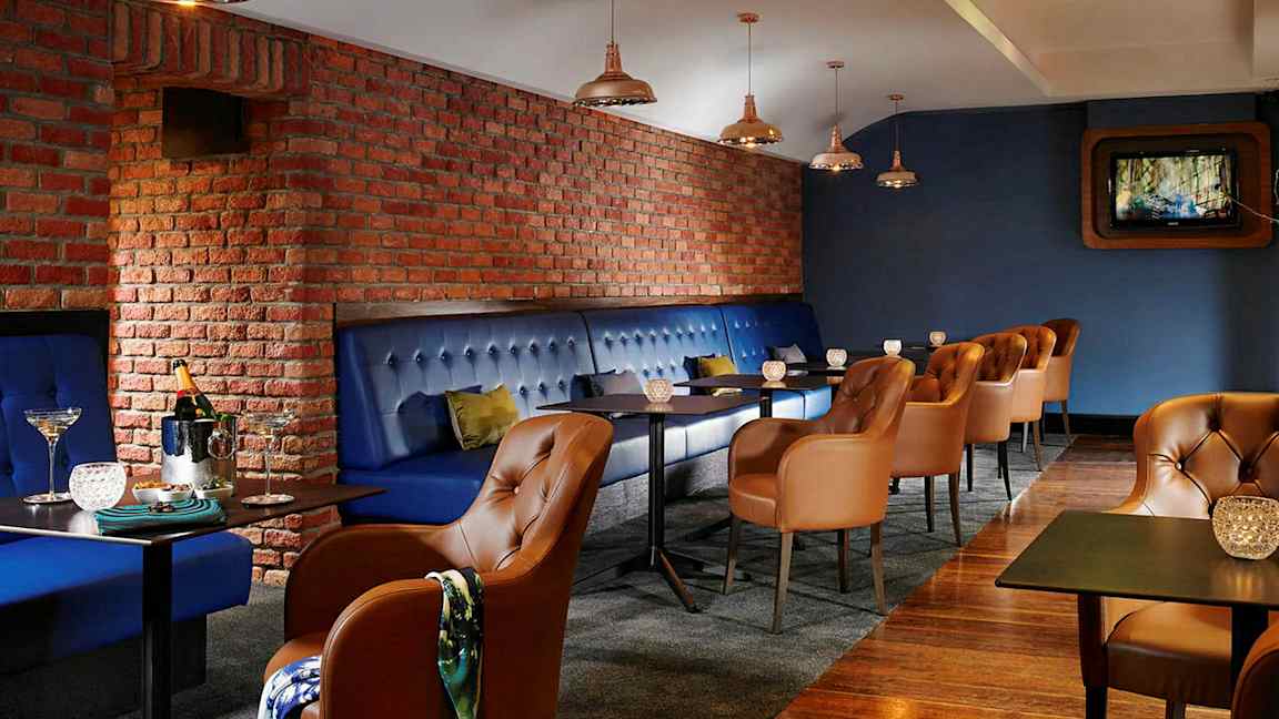 En restaurant eller lounge med blåpolstrede bænke og brune læderstole. Væggene er af røde mursten, og der er små borde med dekorative lys. (AI-genereret tekst)