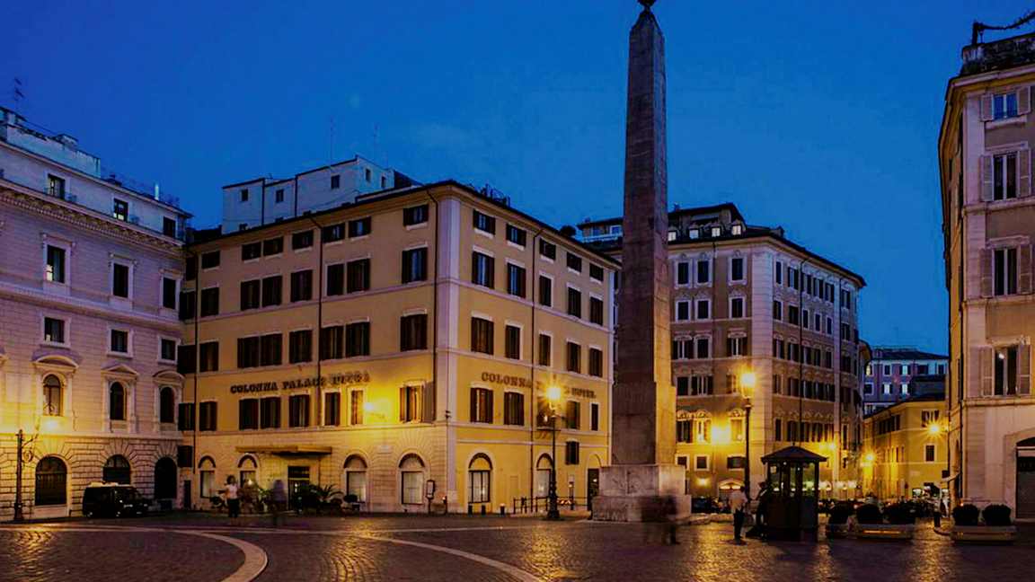 En plads med bygninger omkring og en obelisk i midten, oplyst om natten. (AI-genereret tekst)