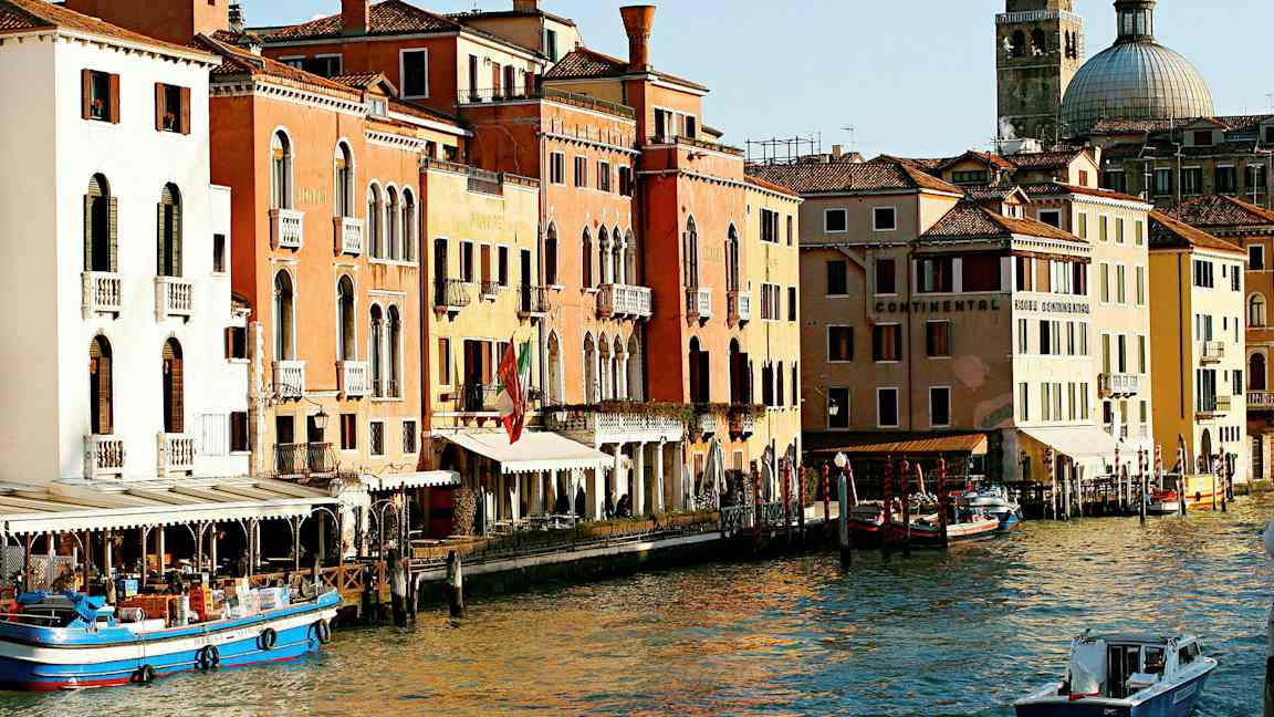 Et billede af en kanal i Venedig med farverige bygninger, både og en kuppelformet kirke i baggrunden. (AI-genereret tekst)