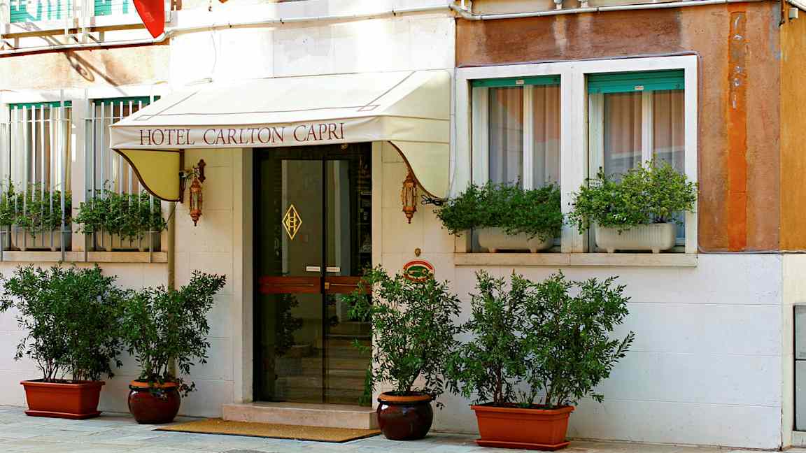 Indgangen til Hotel Carlton Capri med skilt, glasdør og planter. (AI-genereret tekst)