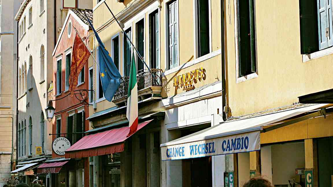 Facader af bygninger med et italiensk flag, europæisk flag og skilte, der angiver hotel og veksel. (AI-genereret tekst)