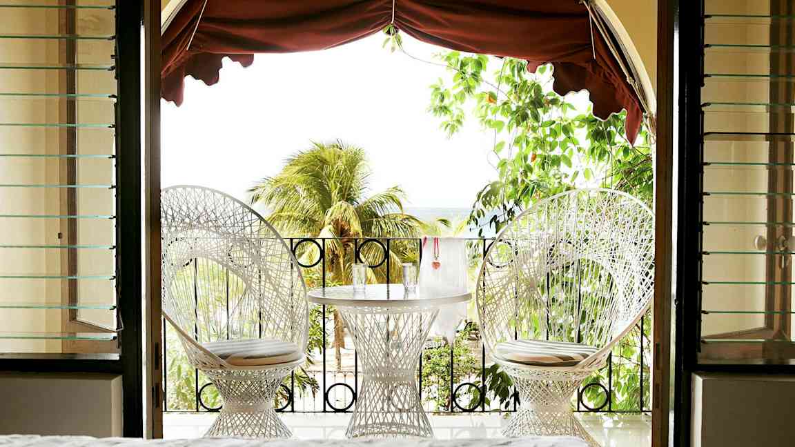 Udsigt fra et rum mod en terrasse med to hvide stole og et bord med glas, omgivet af grønt og palmer. (AI-genereret tekst)