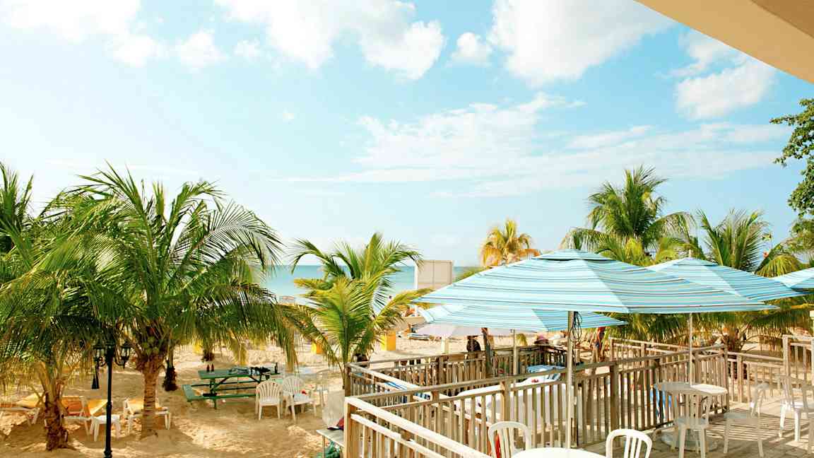 Billede af en strand med palmer, borde og stole samt parasoller. Himlen er klar med nogle skyer. (AI-genereret tekst)