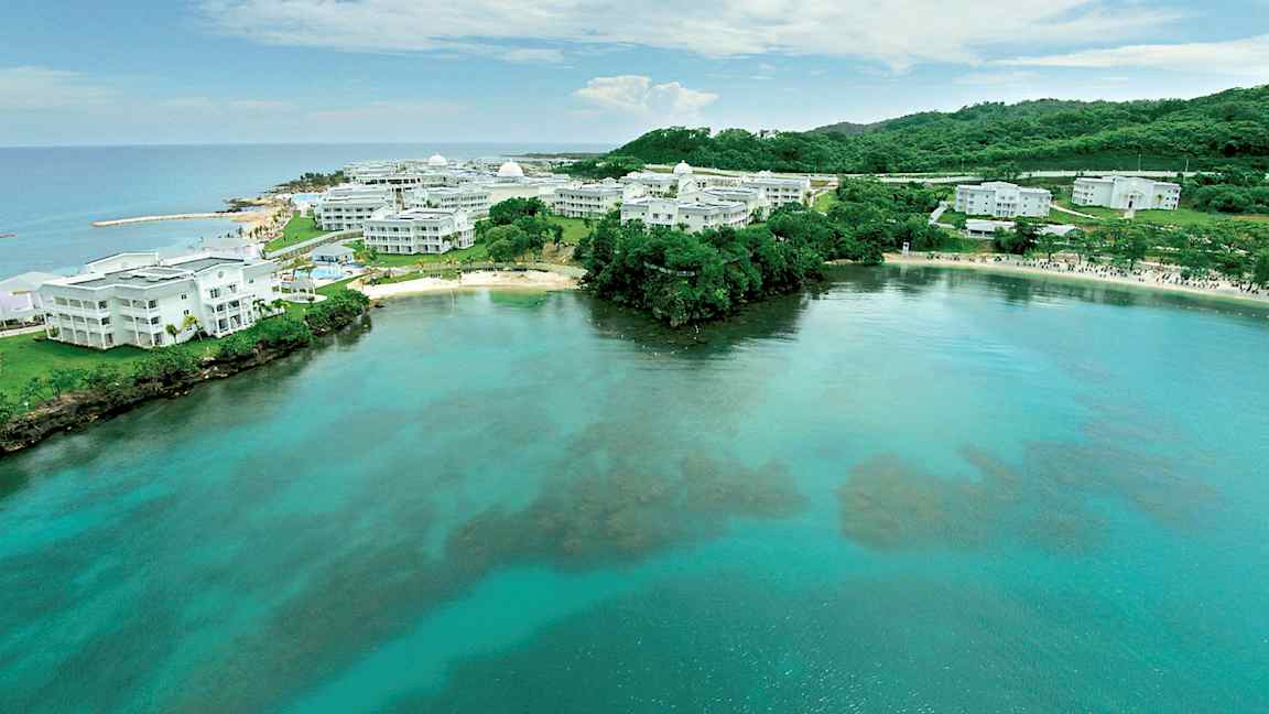 Billede af en kyst med klart vand og hvide bygninger omgivet af grønne områder. (AI-genereret tekst)