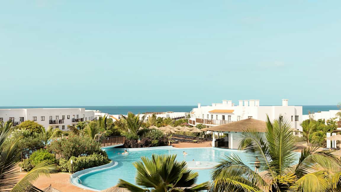 Hotelområde med pool, grønne omgivelser og havet i baggrunden. (AI-genereret tekst)