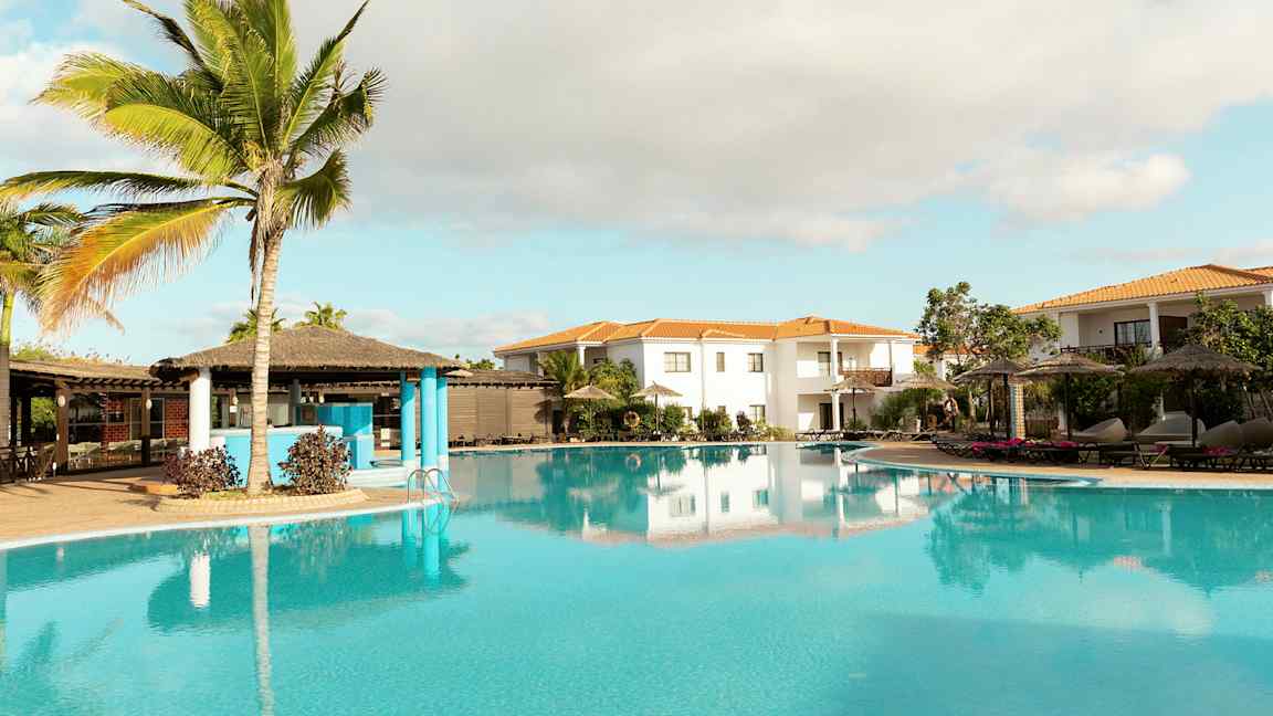 Et tropisk poolområde med palmer, parasoller og omgivende bygninger. (AI-genereret tekst)