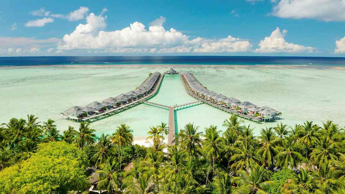 Billedet viser vandbungalower forbundet med gangbroer over en lagune, omgivet af palmer og omgivet af en blå himmel og hav. (AI-genereret tekst)