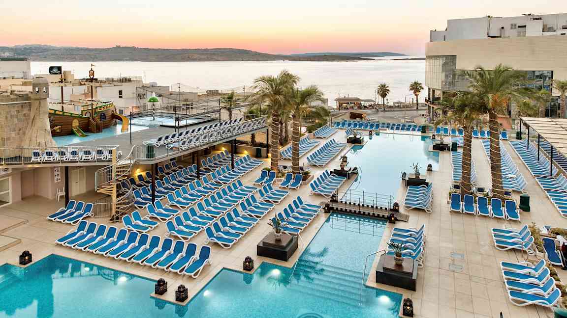 Stort poolområde med liggestole, palmer og udsigt over havet ved solnedgang. (AI-genereret tekst)