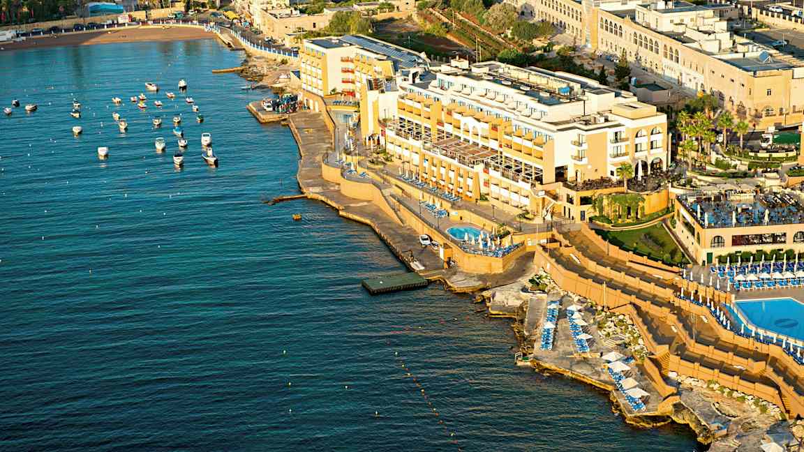 Luftfoto af et kysthotel med pools og liggestole ved havet. Små både er fortøjet i vandet. (AI-genereret tekst)