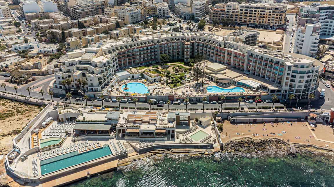 Billedet viser et omfattende feriekompleks med swimmingpools, solområder og omkringliggende bygninger tæt på kysten. (AI-genereret tekst)