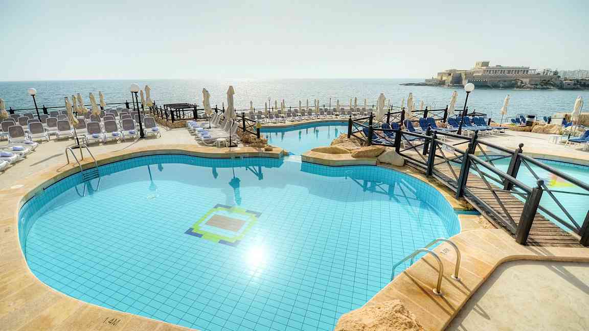 Billede af en udendørs pool ved stranden med liggestole og parasoller omkring. (AI-genereret tekst)