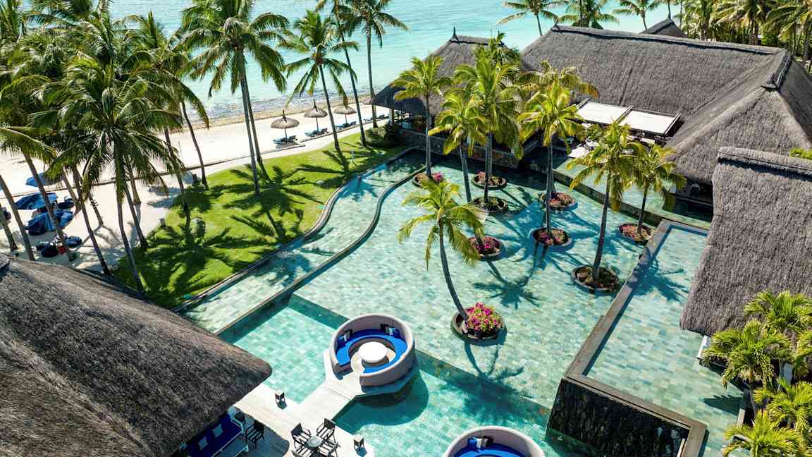 Flere huse med stråtag ved en pool med palmer og sandstrand i baggrunden. (AI-genereret tekst)