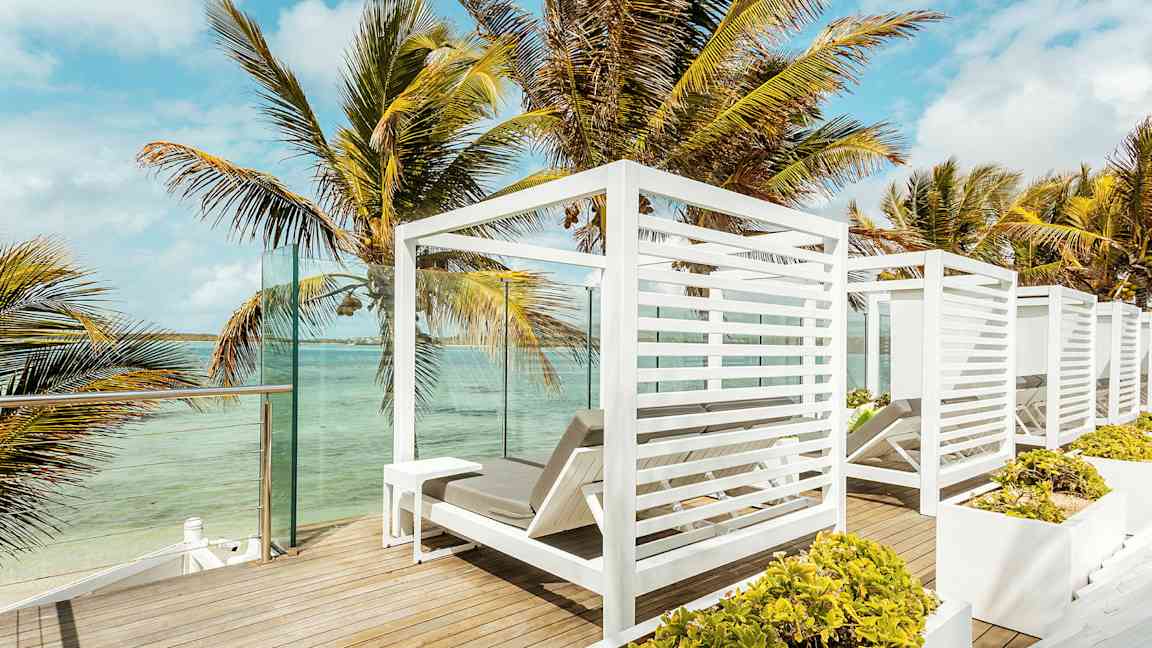 Udsigt over en terrasse med hvide liggestole og palmer ved vandet. (AI-genereret tekst)