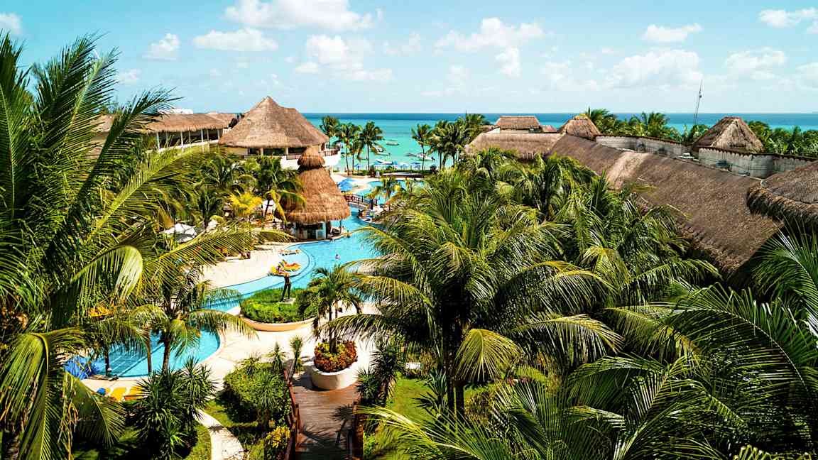 Et stort resort med pools, palmer og bygninger med stråtag, beliggende tæt på havet under en blå himmel. (AI-genereret tekst)