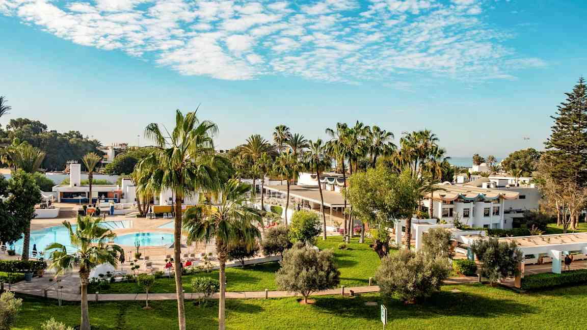 Hotelområde med græsplæner, palmer, pool og hvide bygninger. (AI-genereret tekst)